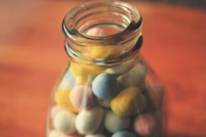 A jar full of pastel coloured sugar almonfs (white, yellow, green .blue,. pink) sitting on a brown desk