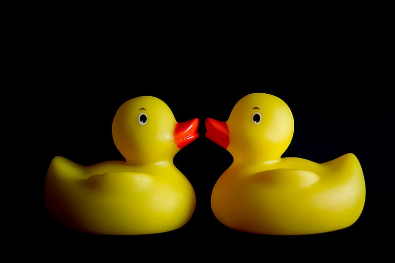 Two yellow rubber ducks with rad beaks looking at each other on a black background