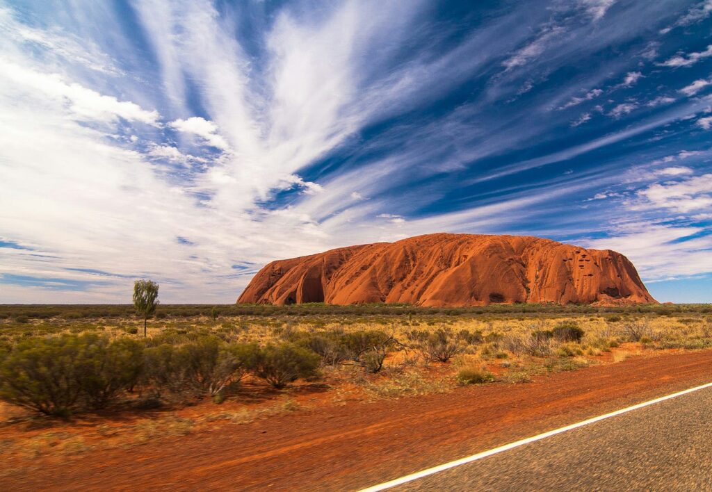 Image of Uluru a large red rock rising from a flat red, saltbush plain with a brillint blue sky