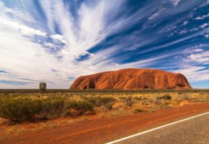 Image of Uluru a large red rock rising from a flat red, saltbush plain with a brillint blue sky